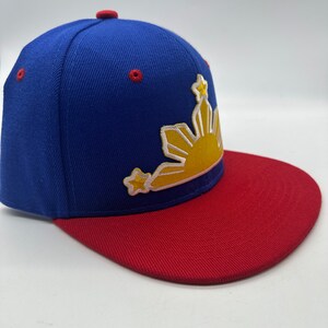 Op de afbeelding: Een blauwe en rode baseballpet met een geborduurd zon- en sterrendesign in geel en wit. De zon heeft een gele omtrek en de sterren zijn wit.