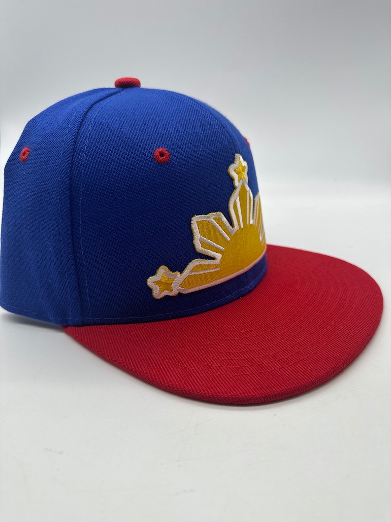 Op de afbeelding: Een blauwe en rode baseballpet met een geborduurd zon- en sterrendesign in geel en wit. De zon heeft een gele omtrek en de sterren zijn wit.