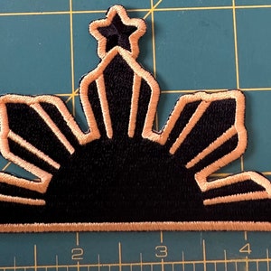 May include: A black and tan embroidered patch featuring the sun with rays and three stars, representing the Philippine flag.
