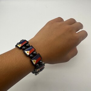 Puede incluir: Un brazalete de cuentas negras con peque&ntilde;os dijes rectangulares que muestran la bandera de Filipinas. Los dijes se alternan con cuentas negras.