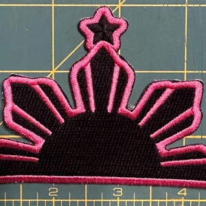 Puede incluir: Parche bordado con un diseño de sol negro con contornos y detalles en rosa. El parche incluye tres estrellas rosas y mide aproximadamente 7.6 cm de ancho. El parche está sobre una esterilla de corte verde.