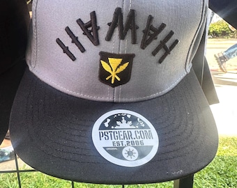 Può includere: Un cappellino da baseball grigio e nero con la scritta "HAWAII" ricamata in nero. Il cappellino presenta un emblema giallo e nero e un adesivo con la scritta "PSTGEAR.COM EST.2006 ORIGINAL".