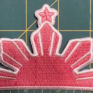 May include: Embroidered patch featuring a stylized sunburst design in shades of pink and white. The patch includes three five-pointed stars and is likely intended for application on clothing or accessories.
