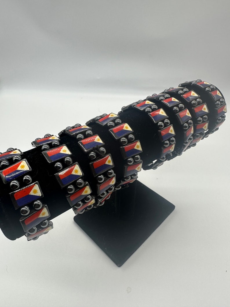 Puede incluir: Una pila de pulseras negras con cuentas plateadas y peque&ntilde;os dijes rectangulares con la bandera de Filipinas. Las pulseras se muestran en un soporte negro.