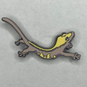 May include: A gecko enamel pin in a relaxed pose. The pin features a light brown body with yellow and purple accents. The gecko has a silver outline and is designed with a cartoonish style.