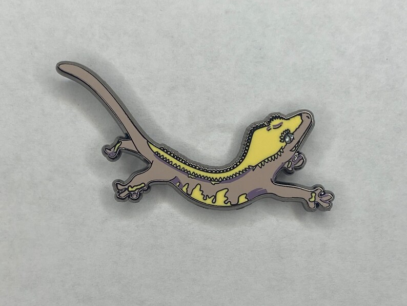 May include: A gecko enamel pin in a relaxed pose. The pin features a light brown body with yellow and purple accents. The gecko has a silver outline and is designed with a cartoonish style.