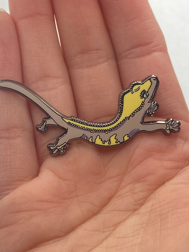 May include: A silver enamel pin depicting a gecko in a running pose. The gecko has a yellow head and a light brown body. The pin is detailed and likely a collectible. The gecko's body is approximately 7 cm long.