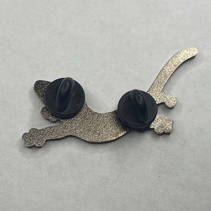 May include: A silver-coloured cat-shaped enamel pin with two black pin backs. The pin has a textured surface and a playful design of a cat in motion. Suitable for jackets, bags, or hats.