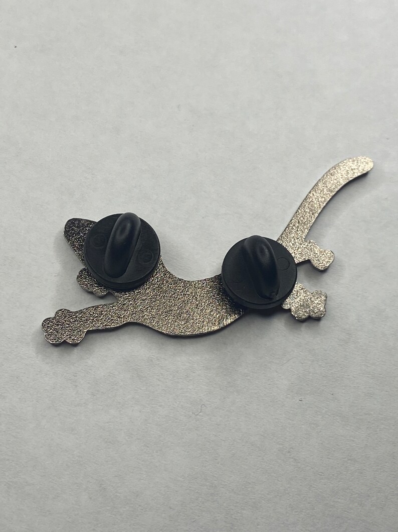 May include: A silver-coloured cat-shaped enamel pin with two black pin backs. The pin has a textured surface and a playful design of a cat in motion. Suitable for jackets, bags, or hats.