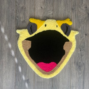 May include: A yellow, black, and pink tufted rug in the shape of a cartoon character's open mouth. The rug features a black mouth, pink tongue, and yellow and brown accents. The rug is placed on a wooden floor.