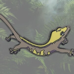 May include: A gecko-shaped enamel pin in yellow and grey. The gecko is shown in a relaxed pose with closed eyes. The pin has a silver outline and is set against a blurred background of green foliage.