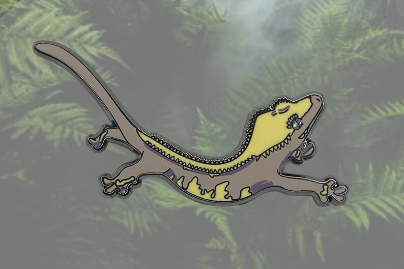 May include: A gecko-shaped enamel pin in yellow and grey. The gecko is shown in a relaxed pose with closed eyes. The pin has a silver outline and is set against a blurred background of green foliage.