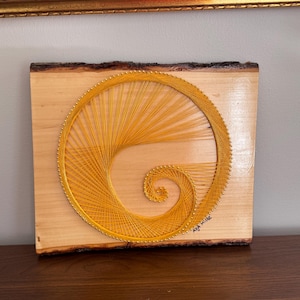 May include: A yellow string art piece in the shape of a spiral on a wooden board. The board has a natural edge and the artist's signature is in the bottom right corner.