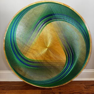 May include: Circular string art piece with a wooden frame. The design features concentric waves of green, blue, and gold threads, creating a swirling pattern. The artwork is mounted on a light-colored wall and rests on a wooden floor.