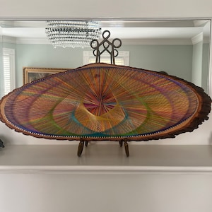 May include: A colorful string art piece on a wooden slice. The art features a circular design with a variety of colors, including red, yellow, green, blue, and purple. The wooden slice is oval-shaped and has a natural bark edge.