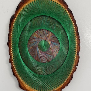 May include: A handcrafted string art piece on a wood slice. The artwork features concentric circles of green thread, with a multicolored center. The wood has a natural bark edge and is adorned with small gold-colored pins. Signed by the artist.