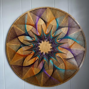 May include: A round wooden wall art piece with a geometric design. The artwork features a central starburst pattern with colorful threads in shades of purple, orange, and teal. The threads are intricately woven, creating a unique and eye-catching design.