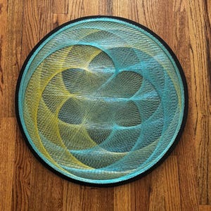 May include: A round string art piece with a black frame. The design features interwoven yellow and teal threads, creating a geometric pattern. The artwork is displayed on a wooden surface.