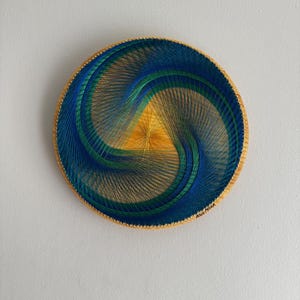 May include: Circular wall art with a spiral design in blue, green, and yellow thread on a wooden base. The artwork has a golden border and is mounted on a white wall. The diameter is approximately 25 cm.