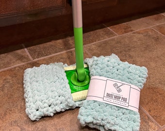 May include: A light green, chunky knit, reusable dust mop pad attached to a green and silver mop handle, with a second identical folded pad wrapped in a white paper band labeled REUSABLE DUST MOP PAD 100% HANDMADE with a line art icon.