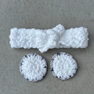 May include: A white, crocheted headband with a knotted detail, paired with two matching round facial pads. The pads have a white center and a black and gray border. The items are arranged on a gray surface.