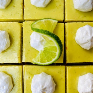 May include: Nine square lemon bars with a white topping and a lime wedge on top of each bar.