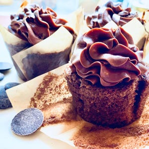 Può includere: Primo piano di cupcake al cioccolato con una ricca glassa al cioccolato a spirale. I cupcake sono in pirottini di carta marrone e alcune scaglie di cioccolato sono sparse intorno. L'immagine ha un'atmosfera calda e invitante.