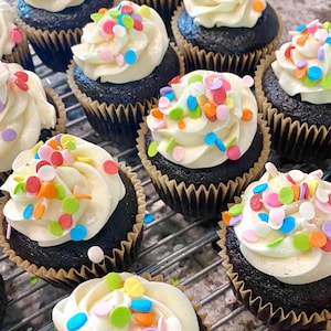 Può includere: Primo piano di una dozzina di cupcake al cioccolato con glassa bianca e codette colorate. I cupcake sono disposti a griglia su una griglia metallica.
