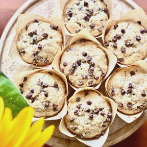 May include: Overhead shot of seven freshly baked muffins with chocolate chips, nestled in brown paper liners, arranged on a wooden serving tray. The muffins are golden brown with scattered chocolate chips on top. A yellow flower is in the lower left corner.