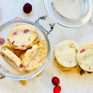 May include: A glass jar filled with white shortbread cookies with red cranberries and pistachios. The cookies are stacked on top of each other. The jar is on a white marble surface with red cranberries scattered around it.