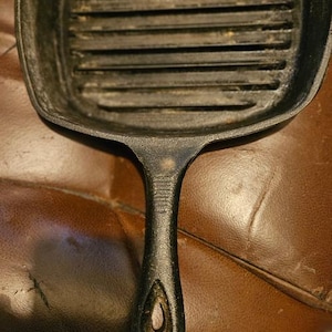 Może przedstawiać: Czarna patelnia grillowa z żeliwa z żebrowaną powierzchnią do gotowania i długim uchwytem. Patelnia ma kwadratowy kształt z zaokrąglonymi rogami i jest przeznaczona do gotowania na płycie kuchennej. Uchwyt ma otwór do zawieszania.