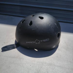 Könnte beinhalten: Ein mattschwarzer Pro-Tec-Helm mit mehreren Belüftungsöffnungen an der Oberseite und an den Seiten. Der Markenname Pro-Tec ist in Weiß auf der Vorderseite aufgedruckt, begleitet von einem kleinen weißen quadratischen Symbol mit dem Buchstaben M.