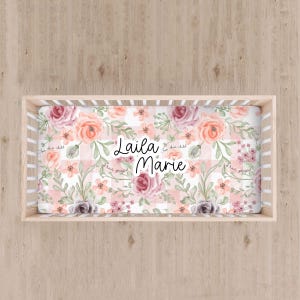May include: A crib sheet with a floral design in shades of pink, peach, and gray. The sheet features the name "Laila Marie" in cursive, with the words "For this child I have prayed" scattered around the edges.