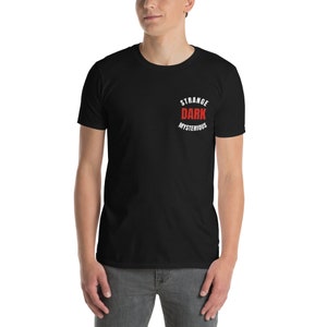 Puede incluir: Una camiseta negra con un gráfico circular blanco que dice "Strange Dark Mysterious" en texto blanco con la palabra "Dark" en rojo.