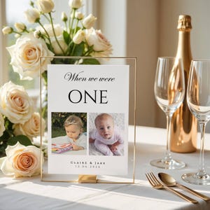 May include: A rectangular gold-framed sign featuring the text "When we were ONE" above two photographs of a baby and the names "CLAIRE & JAKE" with the date "12.06.2026" printed below, displayed on a table with cream roses and wine glasses.