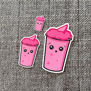 Puede incluir: Tres vasos de batido rosa de dibujos animados con caras sonrientes y ojos saltones. El vaso más grande tiene un remolino de glaseado rosa en la parte superior.