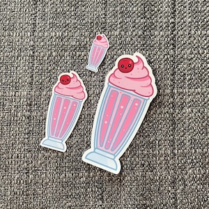 May include: Three pink and white milkshake stickers with a red cherry on top. The largest sticker has a smiling face on the cherry.