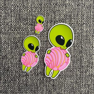 Könnte beinhalten: Drei Aufkleber mit Cartoon-Aliens. Jeder Alien hat einen grünen Kopf mit großen schwarzen Augen und einen rosa-gelb gestreiften Körper. Die Aufkleber befinden sich auf einem strukturierten grauen Hintergrund.