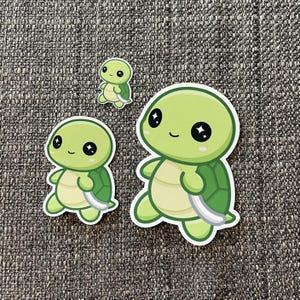 May include: Three green turtle stickers with sparkly eyes. The largest turtle is smiling and has a shell with a green and white pattern. The smallest turtle is standing upright with its legs and arms outstretched.