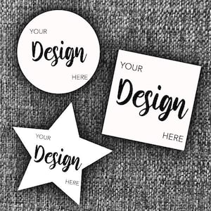 May include: Three white stickers in various shapes: a circle, a square, and a star. Each sticker features the text "YOUR Design HERE" in black script. The stickers are set against a textured grey backdrop.