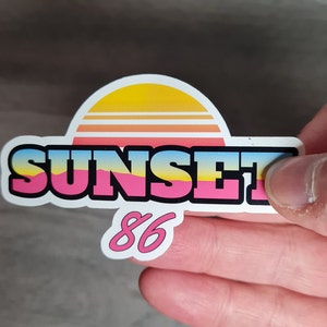 Pu&ograve; includere: Un adesivo bianco con una grafica di tramonto rosa, giallo e blu e il testo "SUNSET 86".