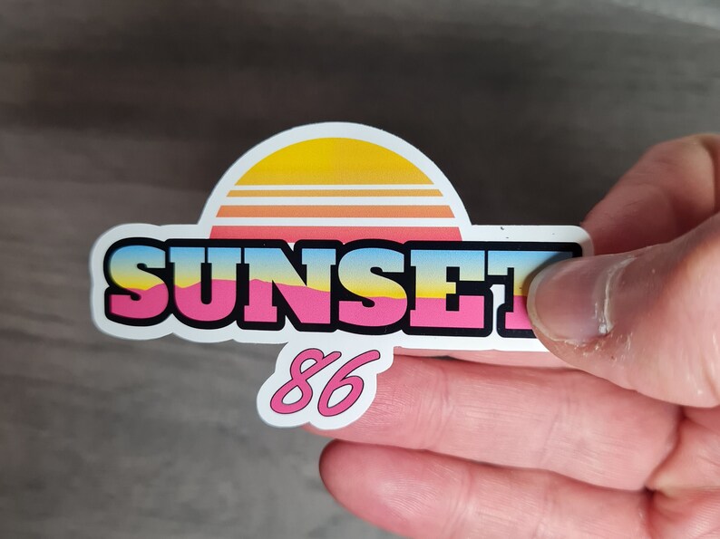 Pu&ograve; includere: Un adesivo bianco con una grafica di tramonto rosa, giallo e blu e il testo "SUNSET 86".