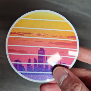 Pu&ograve; includere: Un adesivo rotondo con un design di tramonto retr&ograve; che presenta una silhouette di citt&agrave;. Il tramonto &egrave; rappresentato in tonalit&agrave; di giallo, arancione, rosa e viola, con un contorno bianco. La citt&agrave; &egrave; in tonalit&agrave; di viola e blu.