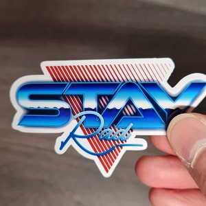 Pu&ograve; includere: Un adesivo bianco con un design blu e rosso. L'adesivo dice "STAY Rad" in un font retr&ograve;.