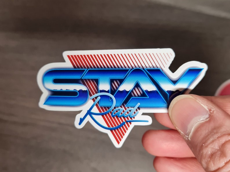 Pu&ograve; includere: Un adesivo bianco con un design blu e rosso. L'adesivo dice "STAY Rad" in un font retr&ograve;.