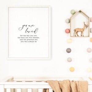 Peut inclure: Une impression encadr&eacute;e blanche avec le texte "you are loved for the boy you are, the man you will become, and the precious son you will always be". L'impression est accroch&eacute;e &agrave; un mur blanc au-dessus d'un berceau blanc avec une couverture marron. Il y a des guirlandes lumineuses avec des boules de coton color&eacute;es accroch&eacute;es au mur.