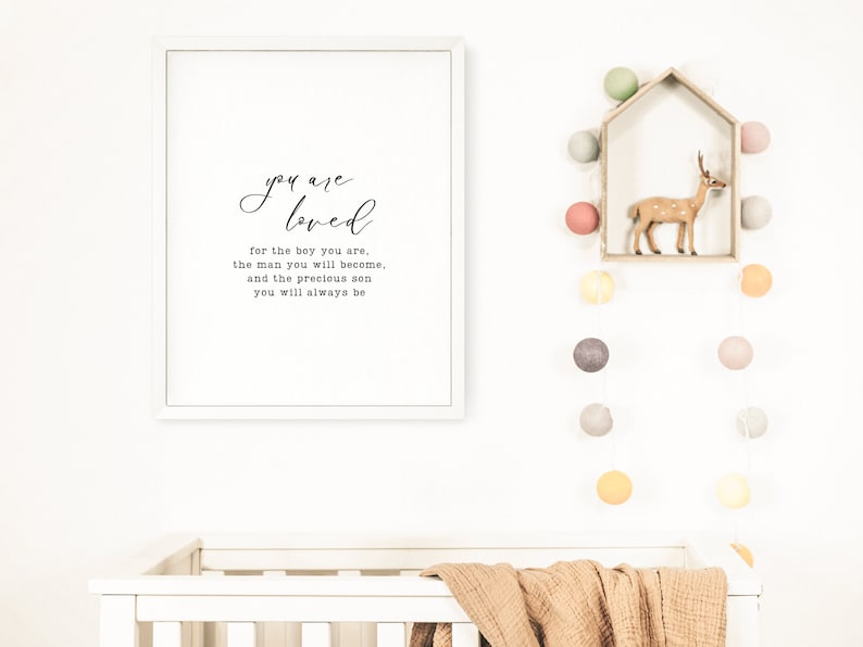 Peut inclure: Une impression encadr&eacute;e blanche avec le texte "you are loved for the boy you are, the man you will become, and the precious son you will always be". L'impression est accroch&eacute;e &agrave; un mur blanc au-dessus d'un berceau blanc avec une couverture marron. Il y a des guirlandes lumineuses avec des boules de coton color&eacute;es accroch&eacute;es au mur.