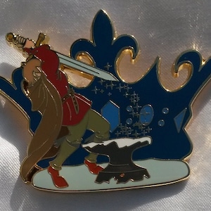 Könnte beinhalten: Eine goldfarbene Anstecknadel, die eine Figur in rot und brauner Kleidung zeigt, die ein Schwert aus einem Amboss zieht. Die Figur steht auf einer weißen Fläche mit einem blauen kronenförmigen Hintergrund. Die Krone hat goldene Akzente und ein blaues und weißes Sternenmuster.