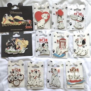 May include: A collection of eleven Disney enamel pins featuring characters from 101 Dalmatians and Cruella de Vil, each mounted on branded cardboard backing cards with paw print designs, showcasing various scenes including puppies, a telephone booth, and text.