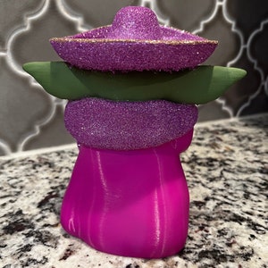Puede incluir: Una figurita de un personaje de color morado y verde que lleva un sombrero morado con ribete dorado. La figurita est&aacute; hecha de un material liso y brillante.